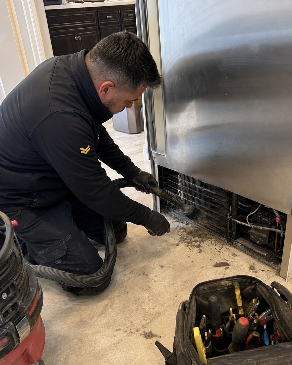 Technician vacuuming and brushing dust from the condenser coils at the base of a built-in Sub-Zero refrigerator