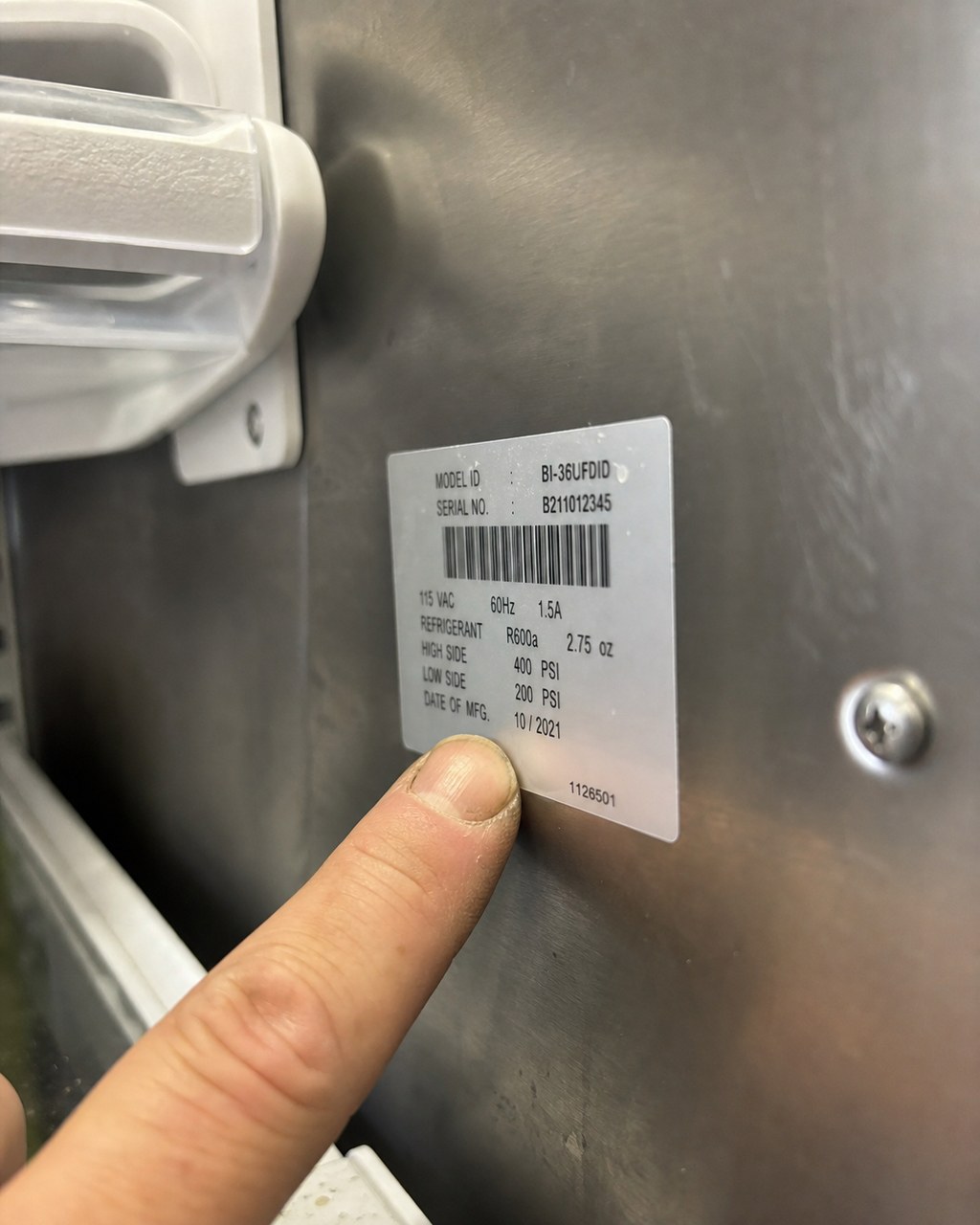 Technician's thumb pointing to the silver model and serial number tag inside a Sub-Zero refrigerator cavity in a Fremont kitchen
